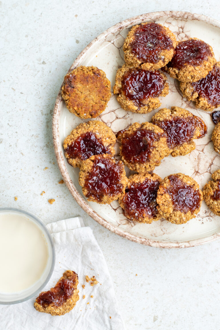 Bovenaanzicht van een beige bord vol met Havermoutkoekjes met pindakaas en aardbeienconfituur, op een beige achtergrond.