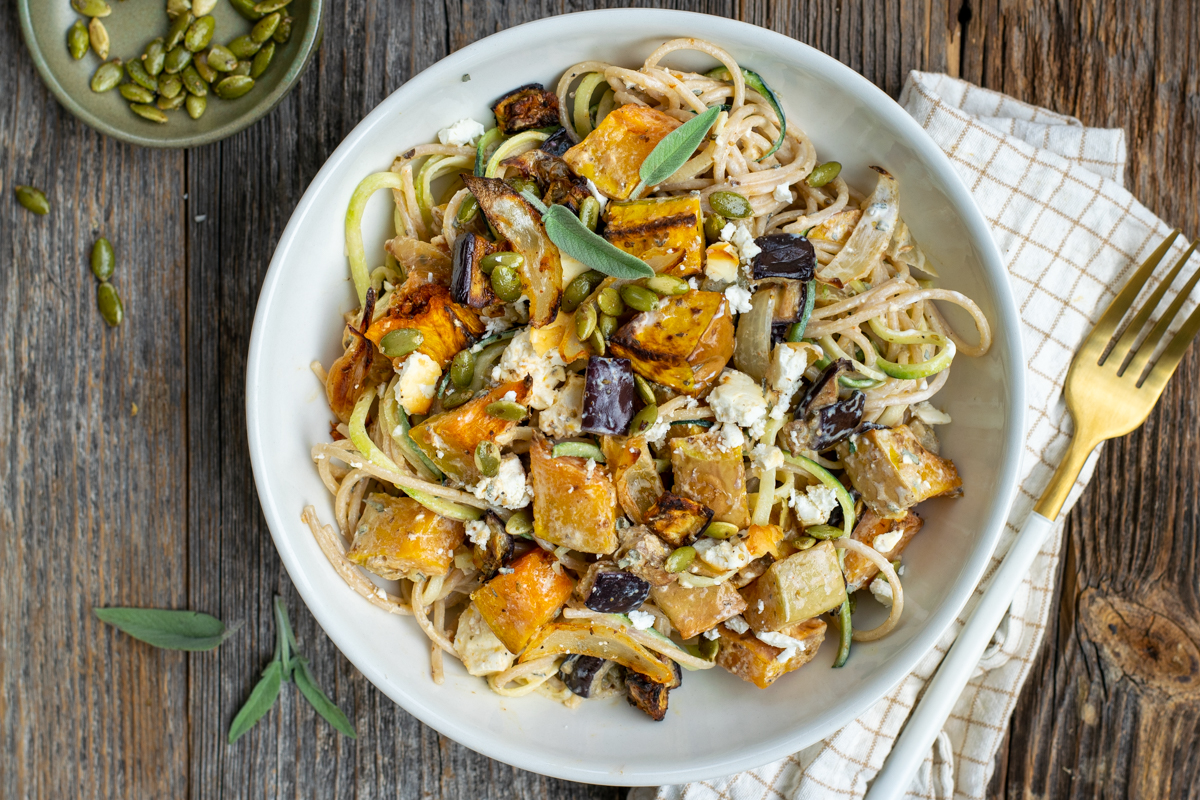 Bovenaanzicht van een wit bord met Romige spaghetti met geroosterde butternut, aubergine en feta; op een ruwe, houten ondergrond.
