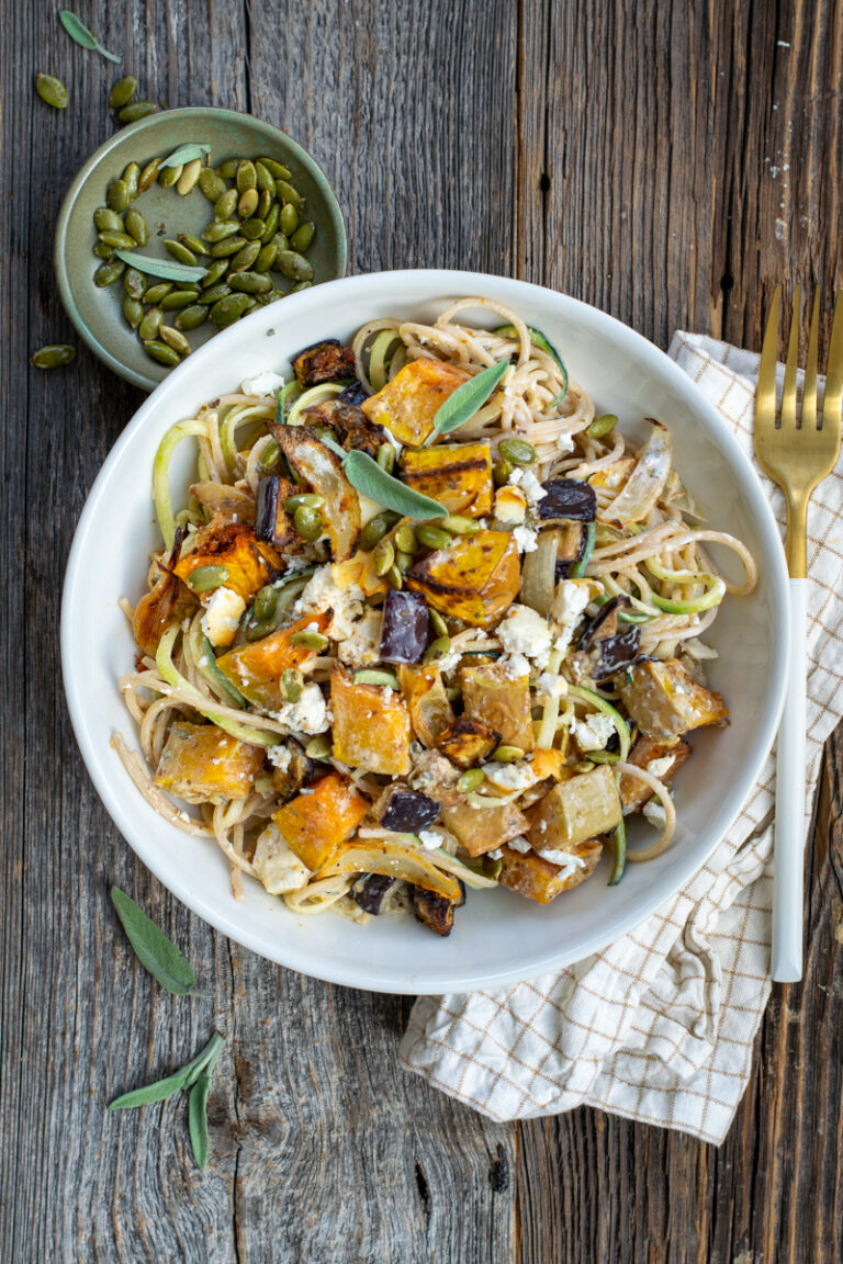 Bovenaanzicht van een wit bord met Romige spaghetti met geroosterde butternut, aubergine en feta; op een ruwe, houten ondergrond.