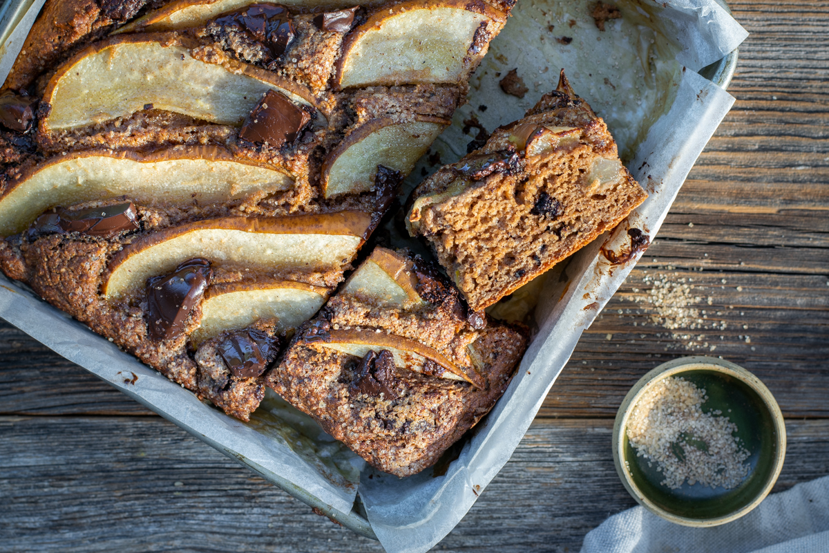 Bovenaanzicht van een vierkante bakvorm op een ruwe houten ondergrond. De vorm bevat een cake met peer en chocolade.