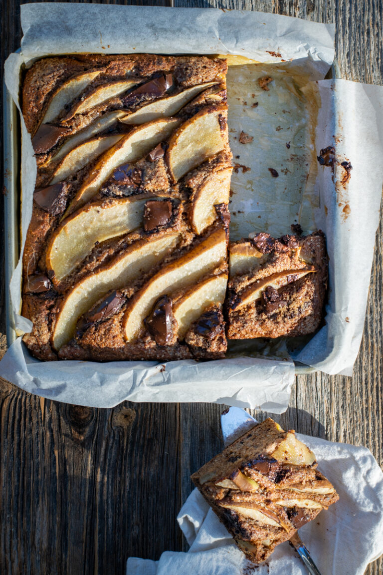 Bovenaanzicht van een vierkante bakvorm op een ruwe houten ondergrond. De vorm bevat een cake met peer en chocolade.