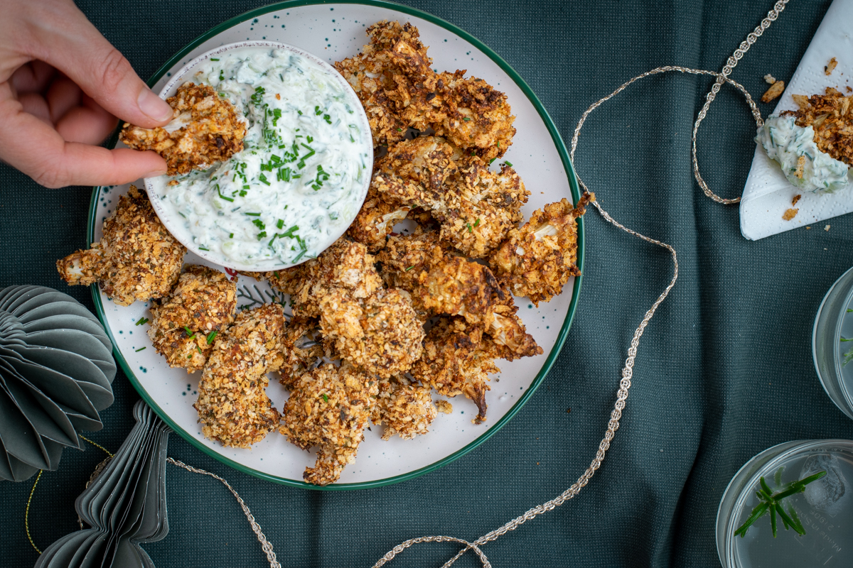 Bovenaanzicht van een bord met knapperige, gepaneerde bloemkoolroosjes, met tzatziki als dip, op een pine groen tafelkleed.