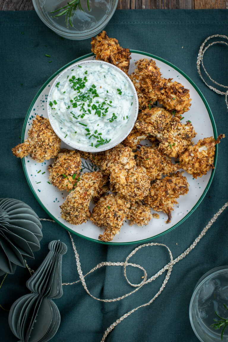 Bovenaanzicht van een bord met knapperige, gepaneerde bloemkoolroosjes, met tzatziki als dip, op een pine groen tafelkleed.