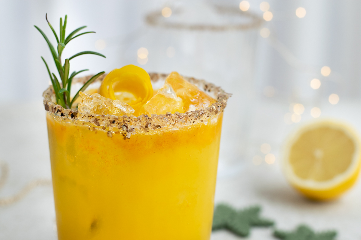 Een goudgele mocktail afgwerkt met rozemarijn en een ruwe rim van zwarte peper en zoutvlokken; op een witte set, met subtiele kerstdecoratie.
