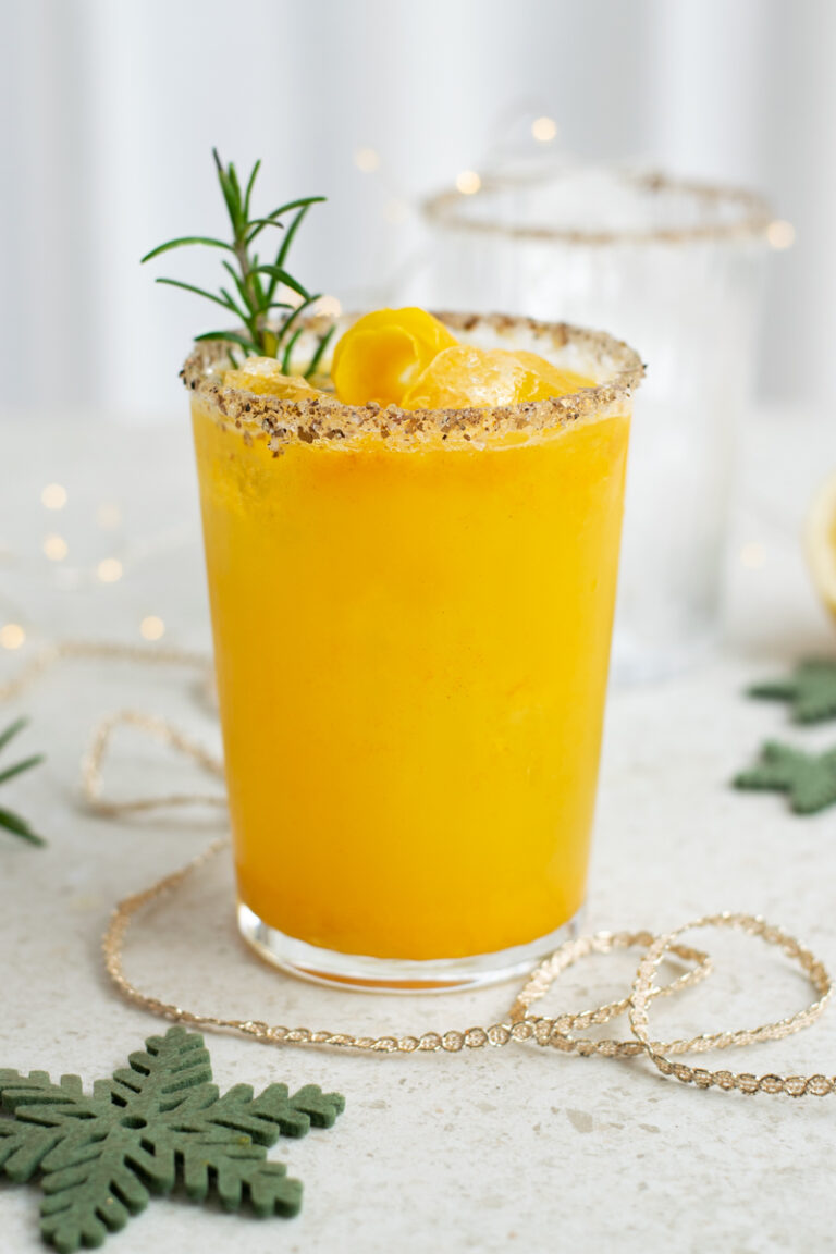 Een goudgele mocktail afgwerkt met rozemarijn en een ruwe rim van zwarte peper en zoutvlokken; op een witte set, met subtiele kerstdecoratie.