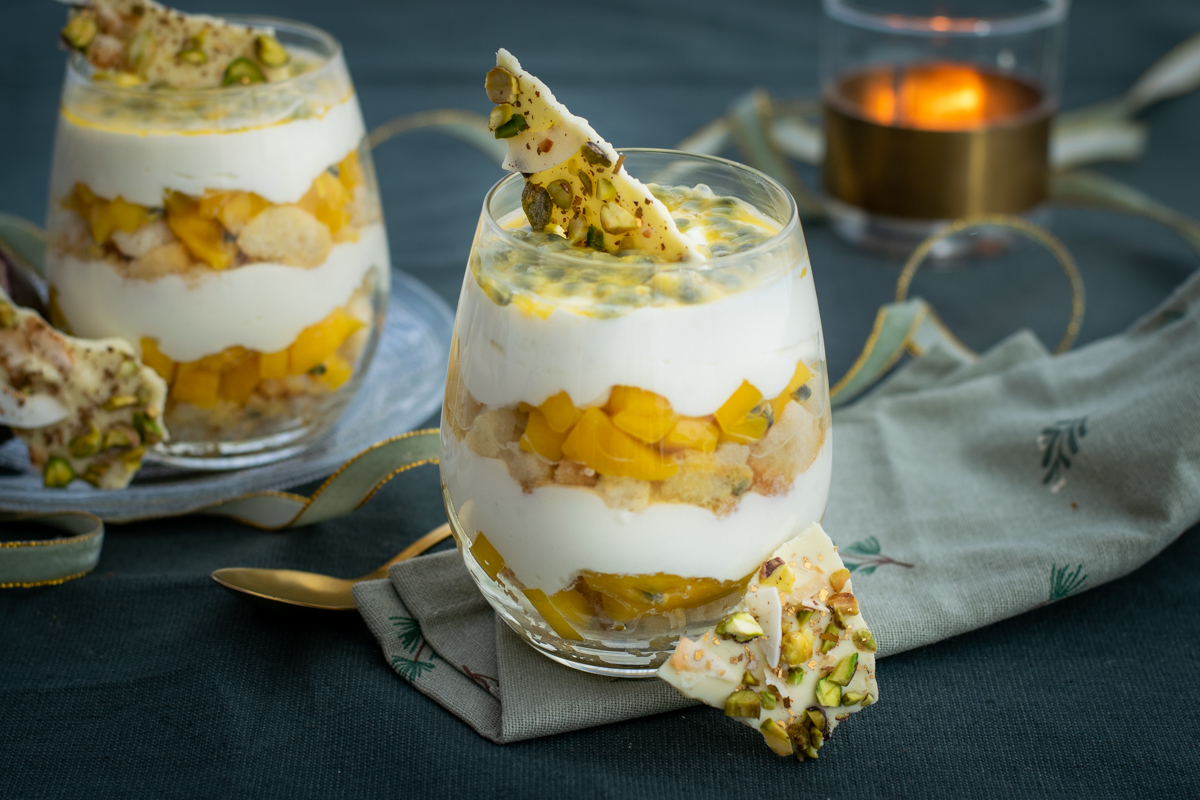 Zijaanzicht van een bol glas, met laagjes boudoirkoekjes, blokjes mango en verse kaas; afgewerkt met een krokantje van witte chocolade en pulp van passievrucht.