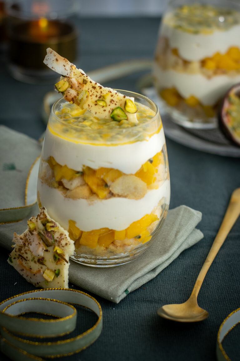 Zijaanzicht van een bol glas, met laagjes boudoirkoekjes, blokjes mango en verse kaas; afgewerkt met een krokantje van witte chocolade en pulp van passievrucht.