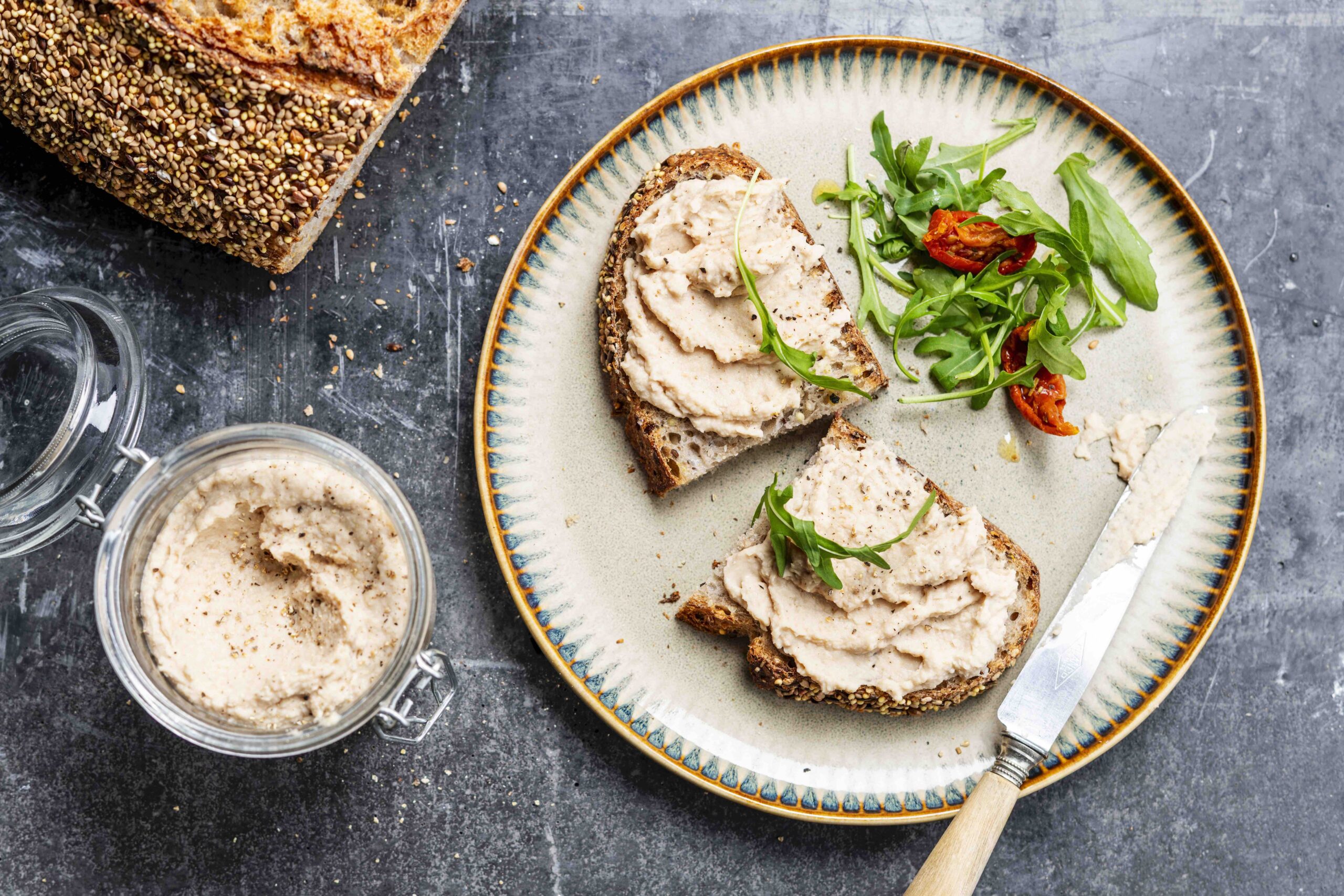 Bovenaanzicht van een snee brood belegd met hummus van witte bonen, afgewerkt met wat rucola, op een donkere ondergrond.