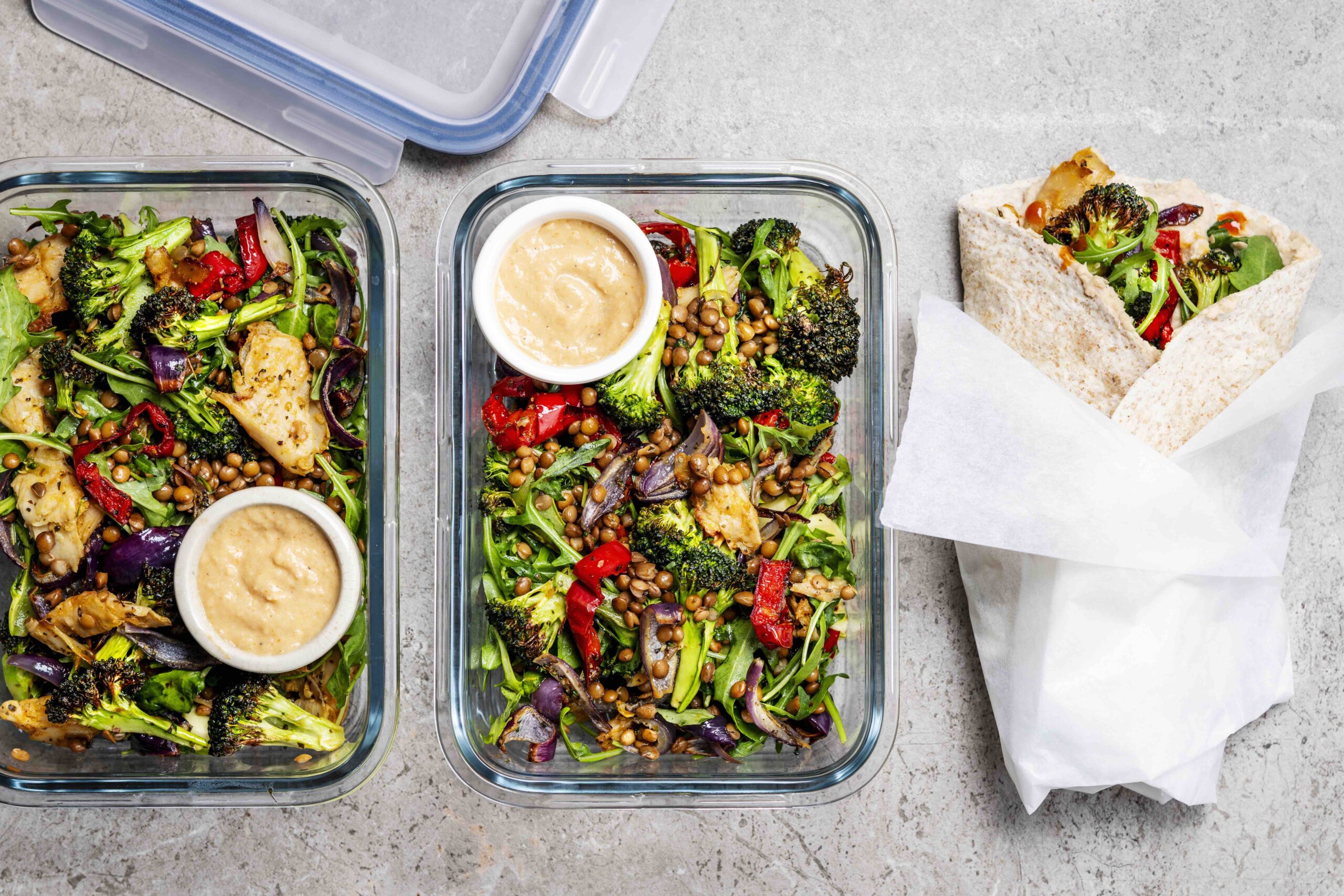 Bovenaanzicht van 2 voorraaddoosjes gevuld met een meal prep salade met geroosterde groenten en kipstukjes en een wrap.
