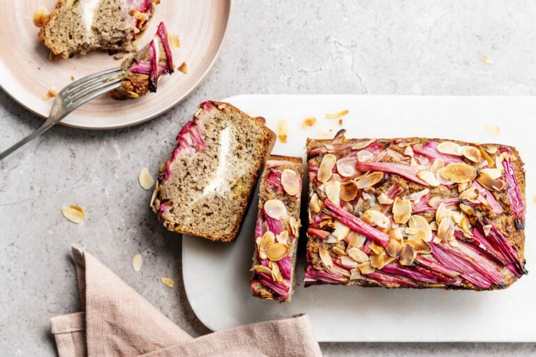 Bovenaanzicht van een rechthoekige rabarbercake met amandel en roomkaas, op een lichtgrijze ondergrond. De bovenkant is afgewerkt met roze rabarberstaafjes en amandelschilfers.