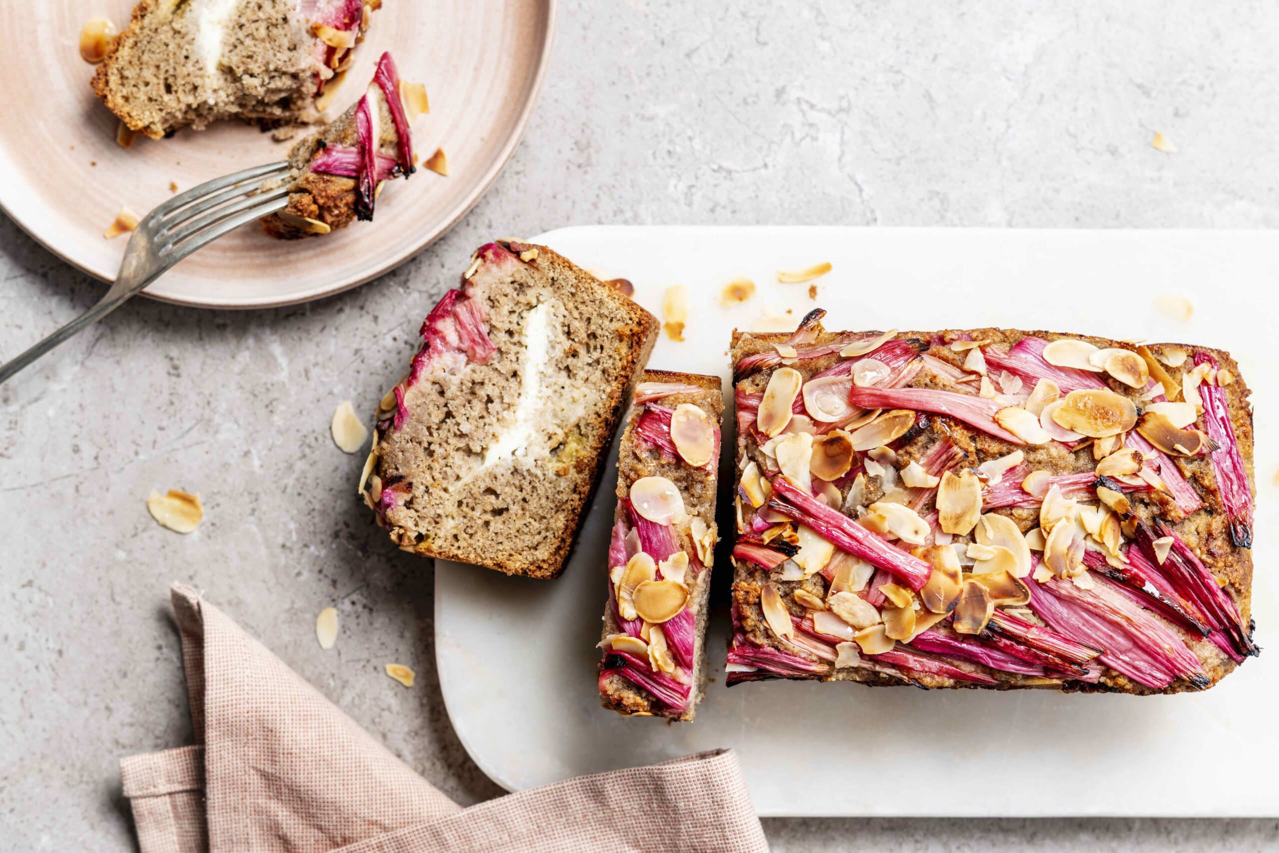 Bovenaanzicht van een rechthoekige rabarbercake met amandel en roomkaas, op een lichtgrijze ondergrond. De bovenkant is afgewerkt met roze rabarberstaafjes en amandelschilfers.